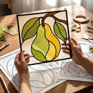 May include: A square stained glass panel featuring a central yellow and green pear design with green leaves and a brown stem, framed in black metal. The panel is held above printed paper patterns labelled Digital Stained Glass Pattern.