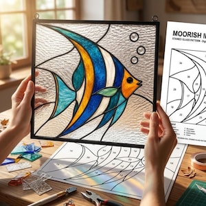 May include: A square stained glass panel featuring a Moorish Idol fish with blue, yellow, and white segments, framed in black metal. Beside it lies a paper pattern labeled MOORISH IDOL STAINED GLASS PATTERN with numbered sections for assembly.