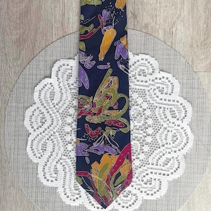 May include: A dark blue necktie featuring an abstract pattern of organic shapes in shades of yellow, purple, green, and magenta. The design consists of scattered, irregular forms with small dot accents distributed across the entire surface of the fabric.