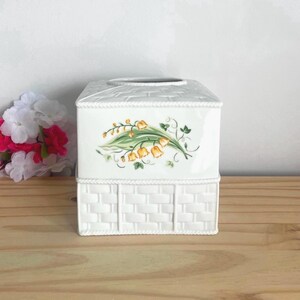 May include: A white ceramic tissue box cover featuring a moulded basket weave texture on the lower section and a smooth upper section decorated with a painted illustration of yellow lily of the valley flowers and green leaves.