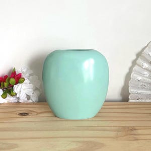 Peut inclure: Un vase en céramique vert menthe, lisse et arrondi, doté d'une large ouverture circulaire sur le dessus. Le vase présente une finition brillante qui reflète la lumière, créant un éclat lumineux sur sa surface incurvée. Un objet décoratif uni.