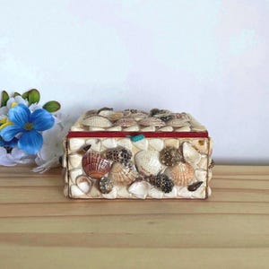 Peut inclure: Une boîte rectangulaire recouverte de divers petits coquillages dans des tons de blanc, beige et marron. Le couvercle présente un fin liseré rouge le long du bord. La surface extérieure est entièrement composée de coquillages collés.