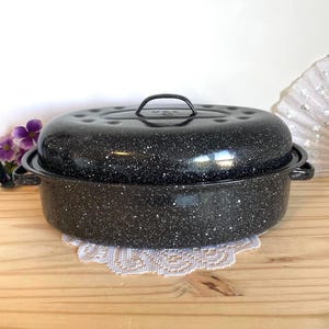 May include: An oval-shaped black roasting pan with a speckled white finish. The pan features two side handles and a matching lid with a central handle. The lid has a series of small indentations along the rim, and the entire piece has a glossy finish.