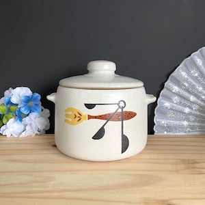 Peut inclure: Un pot en céramique crème avec couvercle et deux anses latérales. La face avant présente un motif peint composé d'une fourchette jaune au manche brun tacheté et de trois cuillères à mesurer noires disposées en groupe sur un fini moucheté.