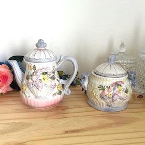 May include: A ceramic teapot and a matching sugar bowl with lids, both featuring a raised relief design of a fruit basket with yellow, pink, and purple accents. The pieces have a white base, light blue trim, and a fluted texture on the lids.