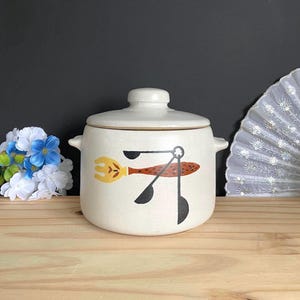May include: A cream-coloured ceramic lidded jar with two side handles. The front features a painted graphic of a yellow fork with a brown speckled handle and three black measuring spoons arranged in a cluster, all set against a speckled finish.