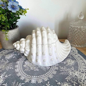 Peut inclure: Un récipient en céramique blanc en forme de coquillage avec un couvercle. Le récipient a une surface texturée avec des crêtes et de petites bosses rondes. Il repose sur un napperon gris avec un motif de dentelle florale blanche. Un pot de fleurs avec des fleurs bleues est en arrière-plan.