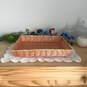 Peut inclure: Un panier rectangulaire en osier rose clair est posé sur une nappe blanche à bords festonnés. Le panier est vide. Le fond comprend des fleurs décoratives et une surface en bois.