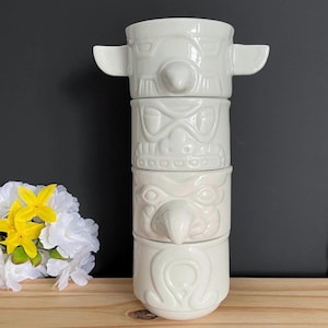 Può includere: Una pila di quattro tazze in ceramica bianca con volti stilizzati in rilievo ed elementi sporgenti. Le tazze sono progettate per incastrarsi verticalmente, creando una struttura continua con tratti del volto e texture variegate.