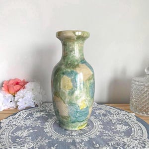 May include: A decorative ceramic vase with a wide base and a narrow neck. The vase is painted with a blend of green, blue, and yellow hues, creating a watercolor effect. The vase is a decorative item.