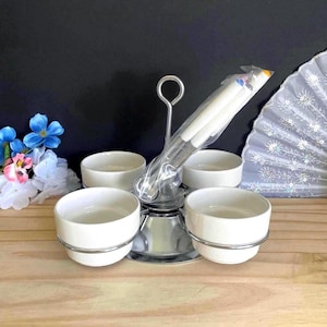 May include: A silver-toned metal serving stand featuring a central handle and four circular holders, each containing a plain white ceramic bowl. A set of metal serving utensils wrapped in clear plastic is positioned upright in the center of the stand.