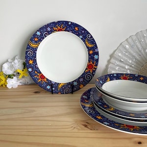 Op de afbeelding: Een set rond keramisch servies met een donkerblauwe rand versierd met gele maansikkels, oranje zonnen en lichtblauwe sterren. Het midden van elk bord en elke kom is effen wit, wat een contrast vormt met het hemelse patroon.