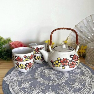 May include: A white ceramic teapot with a brown woven handle and a matching set of three small tea cups. The teapot and cups are decorated with a hand-painted pattern of red and yellow plum blossoms on dark brown branches against a white background.
