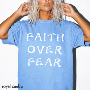 May include: A light blue short-sleeved t-shirt featuring the white text FAITH OVER FEAR printed in a bold, stylised font across the chest. The garment has a crew neckline and a relaxed fit, showcasing a solid colour design throughout the fabric.