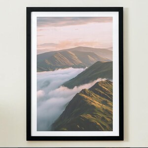 May include: A framed landscape print featuring a mountain range with green, grassy slopes and a valley filled with white clouds. The sky is a soft pink and orange, creating a serene and natural scene. The black frame adds a modern touch.