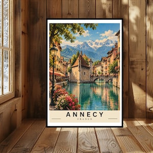 May include: A vertical travel poster featuring a scenic view of Annecy, France, with a stone building on a canal, colourful flowers, a black lamppost, and snow-capped mountains in the background. The text ANNECY FRANCE is printed at the bottom.