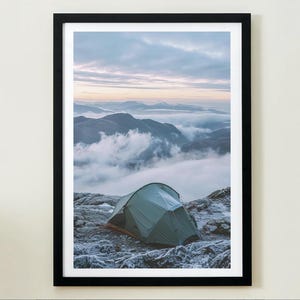 May include: Framed print of a mountain landscape with a tent. A green tent is pitched on a rocky surface, with mountains and clouds in the background. The sky is a blend of blue and white, creating a tranquil and adventurous scene.