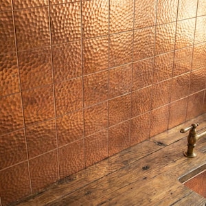 May include: A wall of copper-coloured, hammered-texture tiles, with a wooden countertop and copper sink. A vintage-style brass faucet adds elegance to the kitchen or bathroom. The tiles are approximately 10 cm x 10 cm.