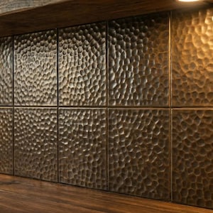 Op de afbeelding: Bronskleurige, getextureerde tegels vormen een muur, verlicht door warme verlichting. De tegels hebben een gehamerd metalen uiterlijk, wat een unieke, rustieke esthetiek creëert. De tegels zijn in een rasterpatroon geïnstalleerd.