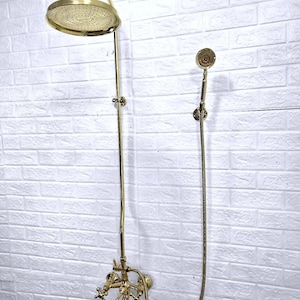 May include: A gold-coloured shower fixture featuring a large, round showerhead and a handheld showerhead. The fixture includes a faucet with cross-shaped handles and a long, curved pipe. The shower is mounted on a white brick wall.