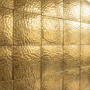 Könnte beinhalten: Eine Wand, die mit quadratischen, gehämmerten Messingfliesen bedeckt ist. Die Fliesen haben eine strukturierte, gewölbte Oberfläche, die das Licht reflektiert und einen warmen, goldenen Farbton erzeugt. Die Fliesen sind in einem Rastermuster angeordnet.