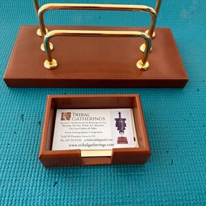 Puede incluir: Un juego de escritorio de cuero marrón que consta de un tarjetero rectangular con tres arcos de metal dorado y un tarjetero cuadrado a juego. El tarjetero contiene una tarjeta de visita blanca de Tribal Gatherings con datos de contacto.