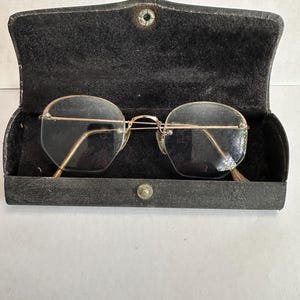 May include: Vintage gold-rimmed eyeglasses with a unique octagonal shape, resting inside a black, textured case. The glasses have a delicate bridge and temples, and the case is secured with a snap closure. The lenses are clear.