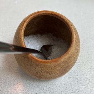 May include: A small, round, brown ceramic salt cellar filled with coarse white salt. A small, silver spoon rests inside the salt. The salt cellar has a rustic, handmade appearance, suitable for a kitchen or dining table.