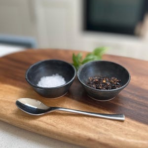 May include: A stainless steel spoon lies on a wooden chopping board beside two small, black ceramic bowls. One bowl holds white salt, the other black pepper. A sprig of green herbs is visible in the background.