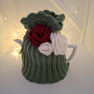 May include: A green crocheted tea cosy with a scalloped edge and three crocheted flowers in red and white. The cosy covers a white teapot. The background is a soft white.