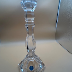May include: A clear crystal candlestick holder featuring a faceted hexagonal base and a slender, contoured stem. The top section has a wide, circular opening for a candle. A small blue circular sticker is attached to the base with text.