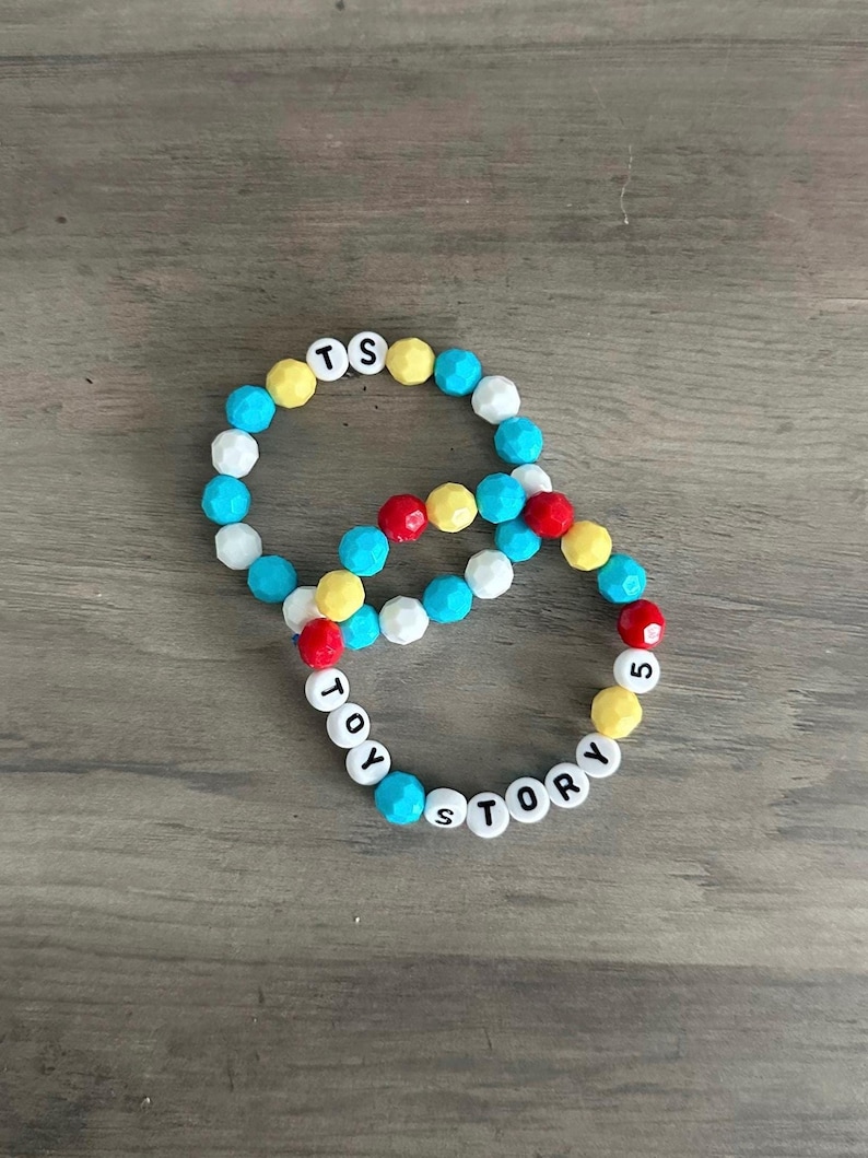 Op de afbeelding: Twee kralenarmbanden met gefacetteerde kralen in de kleuren blauw, geel, wit en rood. E&eacute;n armband bevat witte letterkralen die TS spellen, terwijl de andere de letterkralen TOY STORY 5 bevat, afgewisseld met de kleurrijke kralen.