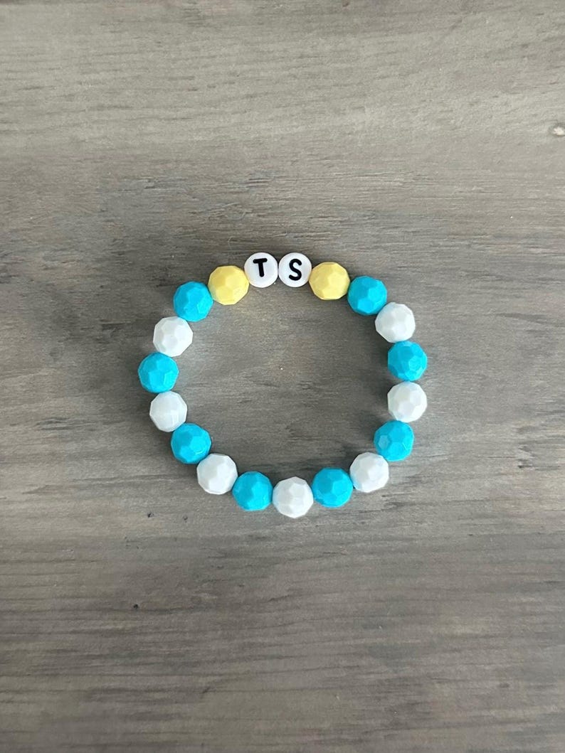 Pu&ograve; includere: Un braccialetto circolare composto da perline sfaccettate in colori alternati turchese brillante, bianco e giallo pallido. Due perline rotonde bianche sono posizionate in alto, ciascuna con una lettera nera, formando il testo TS.