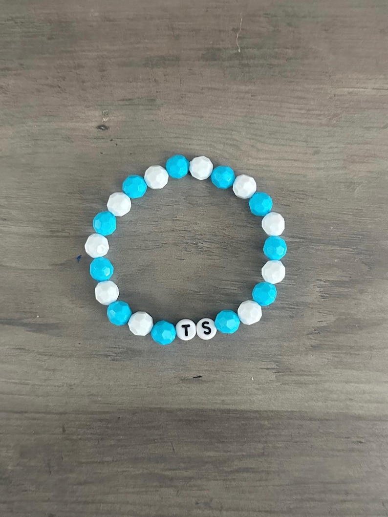 May include: A beaded bracelet with alternating white and turquoise beads. The bracelet features three white beads with the letters "TS" in the center. The bracelet is on a wooden surface.