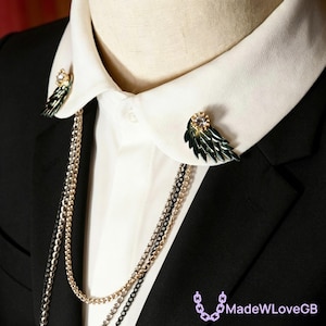 May include: Wing-shaped collar pins with a gold and black chain necklace. The pins are dark green with gold accents and a clear gem. The necklace has gold and black chains. The pins are on a white collared shirt and black suit.