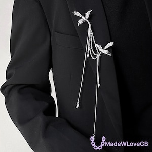 May include: Silver brooch with two bird-shaped ornaments, hanging chains, and pearl details. The brooch is pinned to a black blazer. The design incorporates delicate chains and a small chain at the bottom. The image also includes the text "MadeWLoveGB".