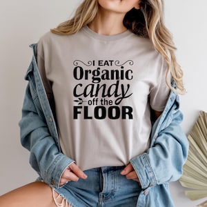 Könnte beinhalten: Hellgraues T-Shirt mit dem schwarzen Text "I EAT Organic candy off the FLOOR". Das T-Shirt mit Rundhalsausschnitt wird mit einer Jeansjacke getragen, was einen lässigen Look ergibt.