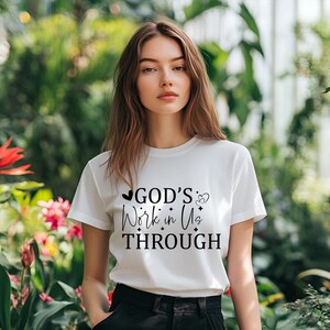 Könnte beinhalten: Weißes T-Shirt mit dem Text "GOD'S Work in Us THROUGH" in schwarzer Schrift. Das Shirt hat einen Rundhalsausschnitt und kurze Ärmel. Der Hintergrund zeigt üppiges grünes Laub und rosa Blumen.