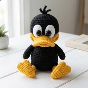 Könnte beinhalten: Ein handgefertigtes gehäkeltes Entenspielzeug in Schwarz und Gelb mit großen weißen Augen und einem gelben Schnabel. Die Ente hat gelbe Füße und sitzt auf einer weißen Holzoberfläche. Eine kleine Topfpflanze und ein Buch sind im Hintergrund zu sehen.