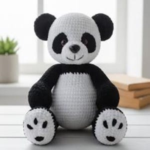 Puede incluir: Un oso panda tejido a mano con hilo negro y blanco. El panda tiene orejas, ojos, brazos y pies negros, con un cuerpo blanco. Está sentado sobre una superficie blanca, con una ventana y una pequeña planta en el fondo.