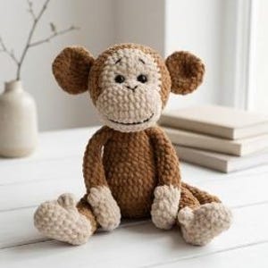 May include: A crocheted monkey toy, crafted from brown and beige yarn. The monkey features large ears and a smiling expression, sitting with its legs extended. A vase with a branch and a stack of books are in the background.