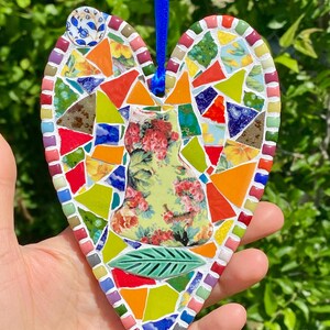 May include: Heart-shaped mosaic ornament with a blue ribbon for hanging. The mosaic features a floral design, a green leaf, and colorful glass pieces in various shapes and sizes. The ornament is held in a hand against a backdrop of green foliage.