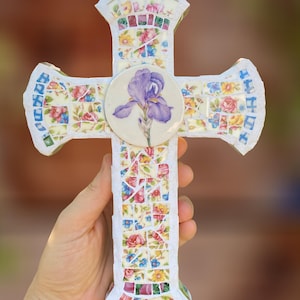 May include: A decorative cross featuring a mosaic design made of small, colorful floral ceramic pieces in shades of pink, yellow, and blue. A circular white ceramic center piece displays a detailed illustration of a single purple iris flower.