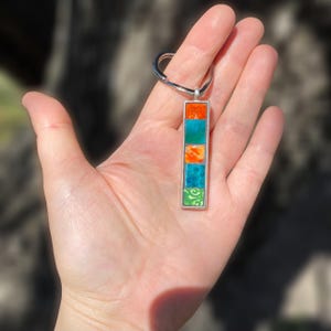 Könnte beinhalten: Ein rechteckiger silberner Schlüsselanhänger-Anhänger mit fünf farbigen Quadraten: orange, türkis, orange, türkis und grün. Das grüne Quadrat hat ein weißes Wirbeldesign. Der Schlüsselanhänger wird von einem silbernen Ring gehalten.