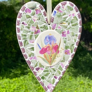 May include: A heart-shaped hanging ornament featuring a mosaic of green and purple ceramic shards surrounding a central oval tile painted with a blue and pink iris flower. The piece is suspended by a light-colored ribbon attached at the top.