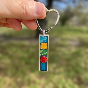 Puede incluir: Un llavero rectangular plateado con un diseño de mosaico colorido. El mosaico presenta cuadrados azules, amarillos, verdes y rojos. El llavero tiene un cierre en forma de corazón.