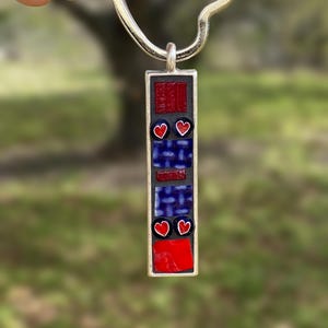 Può includere: Un ciondolo portachiavi rettangolare argentato con un design a mosaico. Il ciondolo presenta tessere di vetro rosse e blu, con due cuori rossi. Il portachiavi è tenuto in mano su uno sfondo verde sfocato.