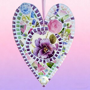 May include: Heart-shaped mosaic art piece with a floral design. The mosaic is made of various pieces of broken china in shades of purple, pink, green, and white. A large purple flower is in the center. A pink ribbon is attached for hanging.