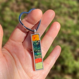 May include: A silver-toned pendant with a rectangular, multi-colored design. The pendant features a heart-shaped bail and a vertical arrangement of orange, green, and red sections. The pendant is held in a hand against a blurred green background.