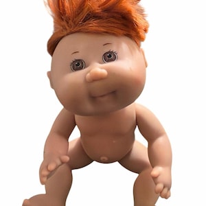 May include: A vintage Cabbage Patch Kid doll with a fair skin tone and red hair. The doll is in a crouching position, with its arms and legs bent. The doll has brown eyes and a small nose. The doll is a collectible item.